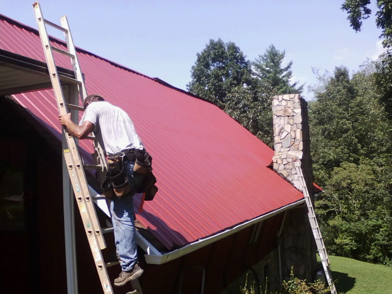 Roofer on a ladder beside a red metal roof and stone chimney — Soffit & Fascia Repair by Roofing Brothers in Alpine